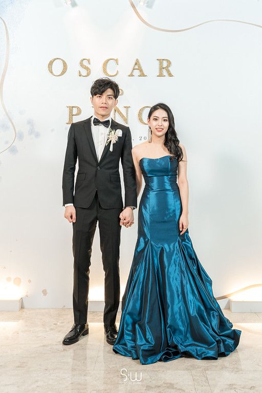A couple poses formally in front of a white backdrop, with the woman wearing a stunning blue mermaid gown.