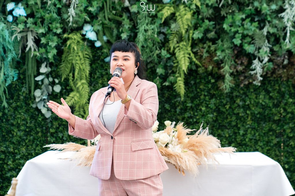 A speaker in a pink suit delivers remarks against a lush green wall with pampas grass decor.