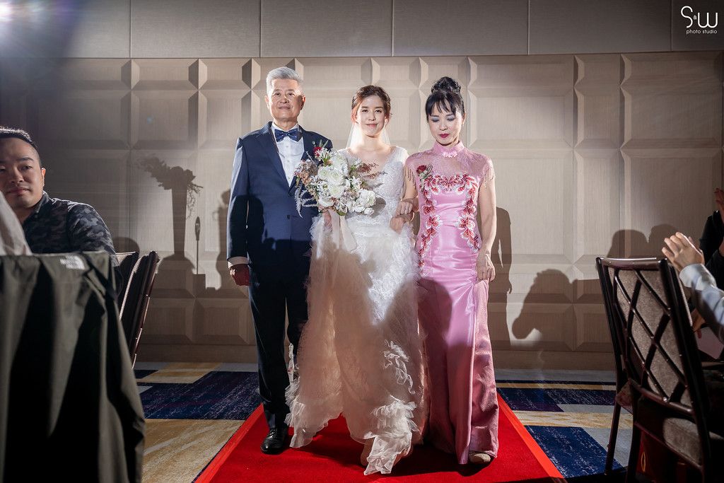 Wedding party members walk down a red carpet in formal attire during a ceremony.