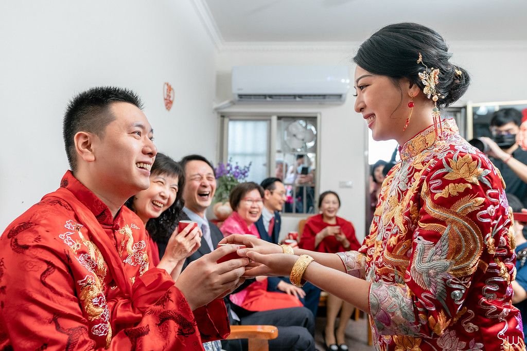 Joyful exchange during Chinese wedding tea ceremony with guests laughing in background.