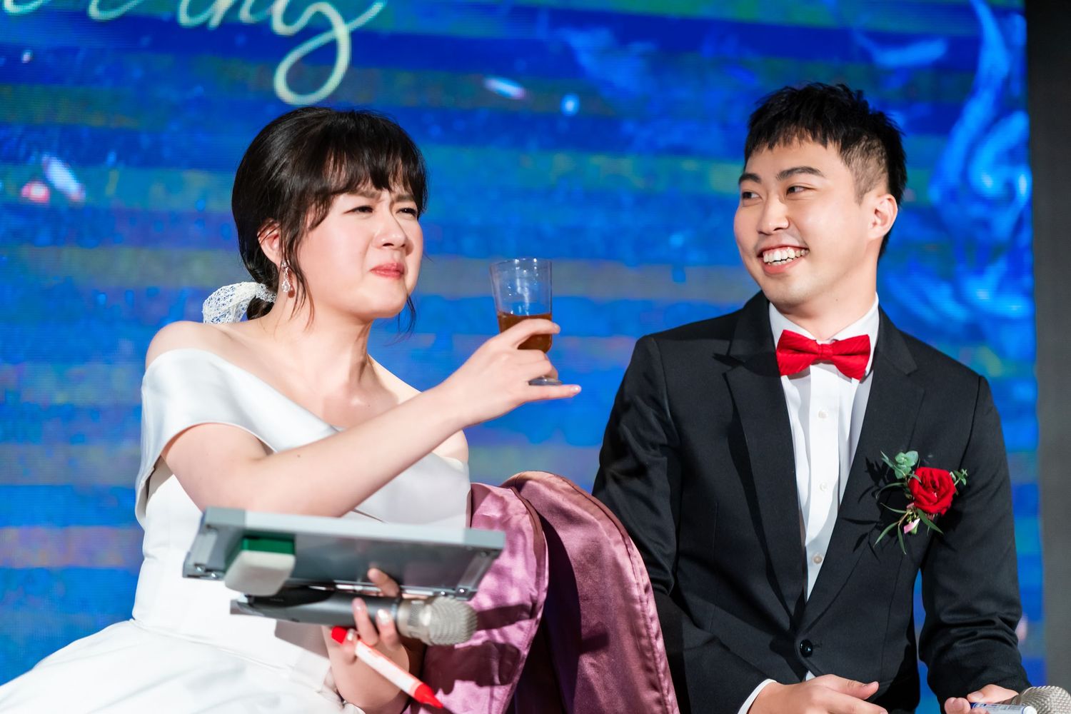 A couple shares drinks during their wedding reception game against a blue backdrop.
