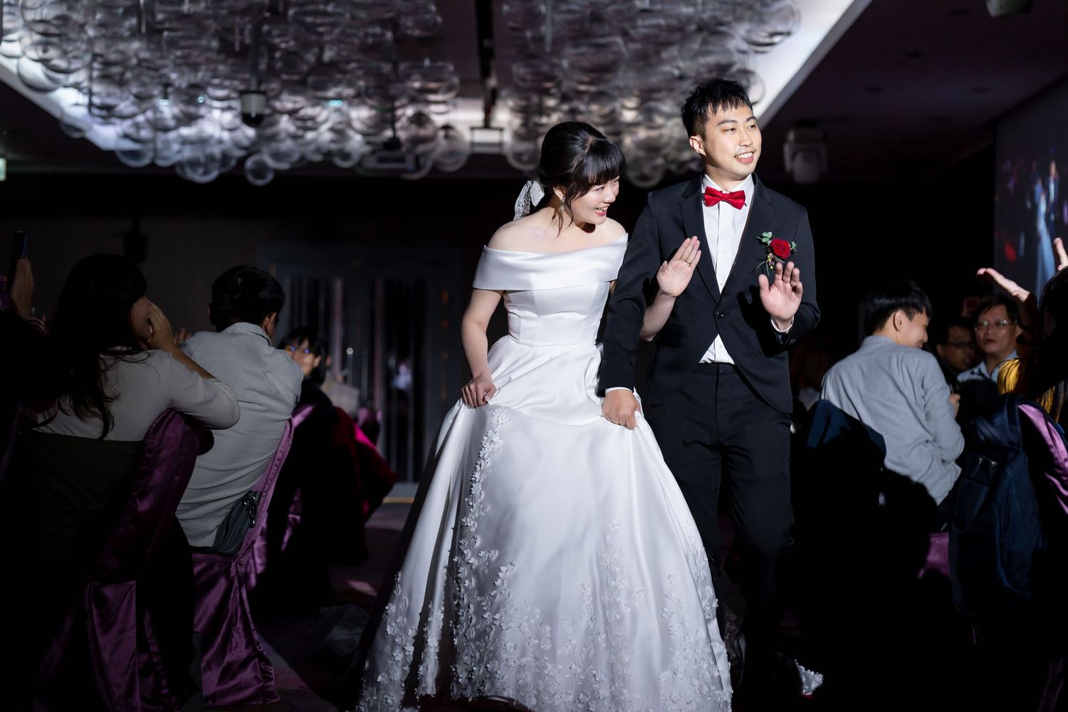 Wedding reception scene with couple dressed in formal attire under dramatic ceiling lighting.