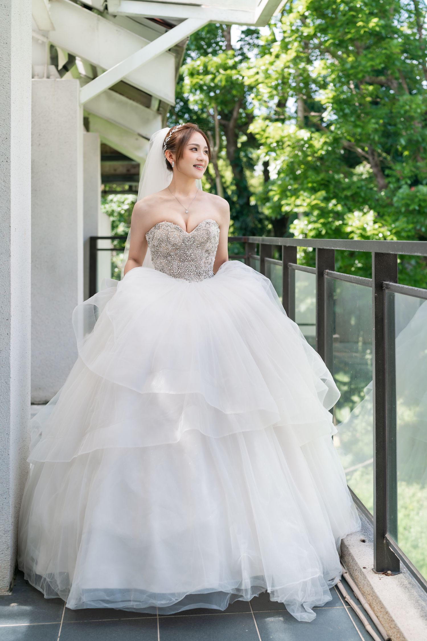 Bride wearing crystal-embellished ballgown poses gracefully on outdoor terrace with glass railing.