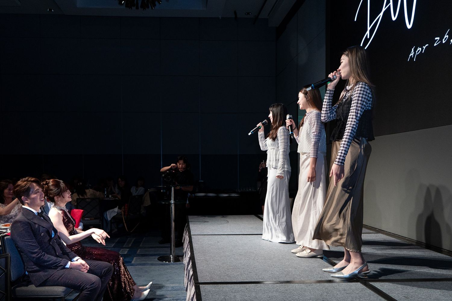 Four women singing on stage with microphones at a formal event, performing for a seated audience in a darkened venue.