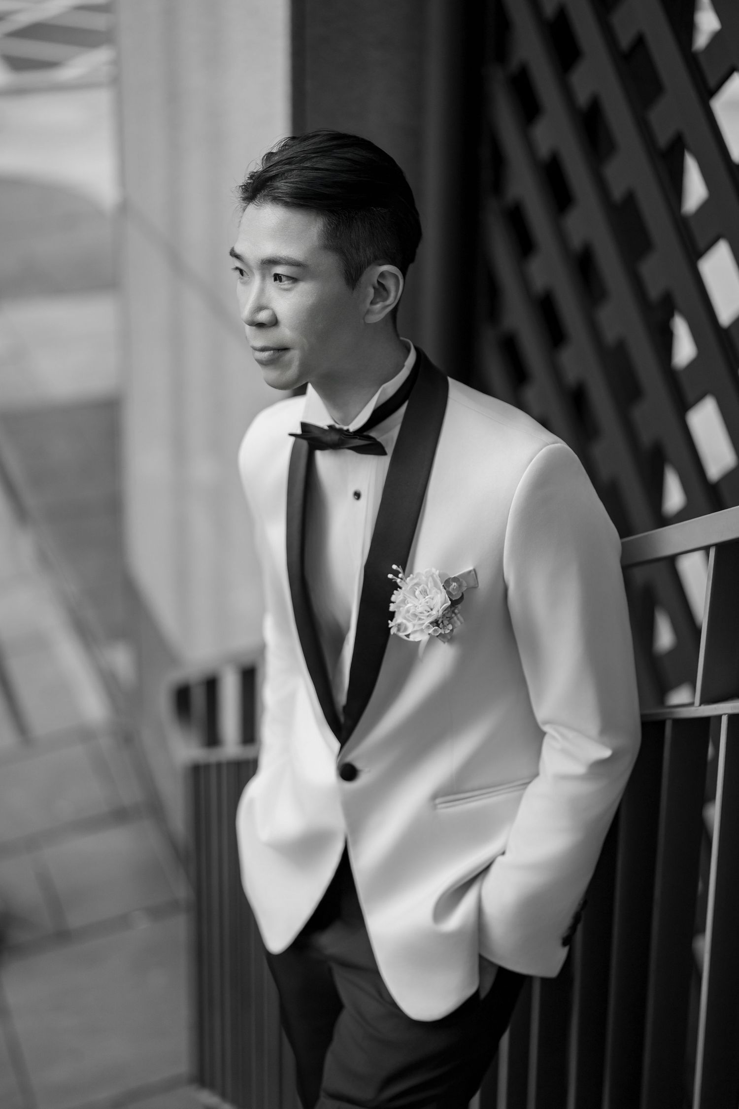 A groom in a white tuxedo jacket with black lapels and bowtie poses against a lattice wall at a formal event.