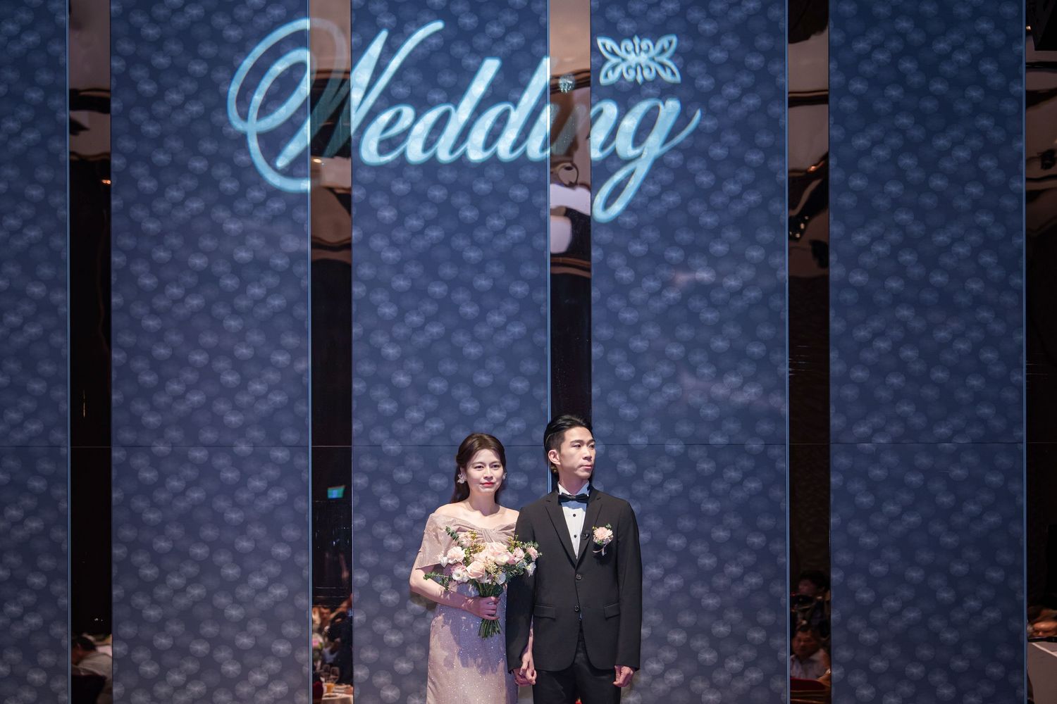 Models walk down a fashion runway wearing elegant black formal attire against a dark blue backdrop with wedding signage.