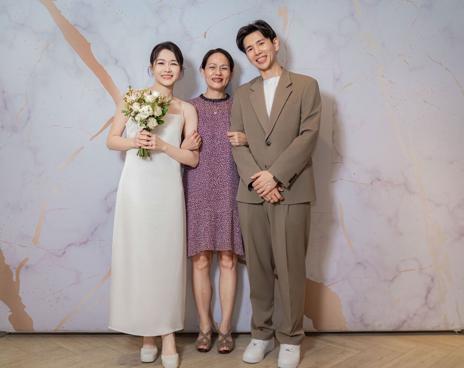 Three people stand together against a marble textured wall in formal attire for photos.