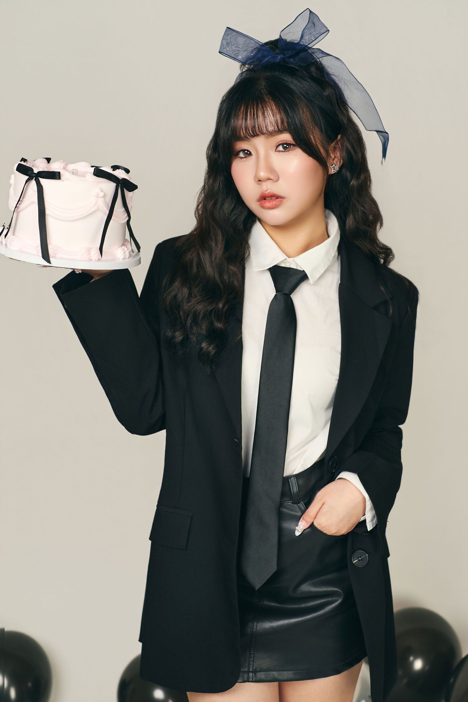 A person in a black blazer and tie holds up a white handbag while posing against a light colored background.