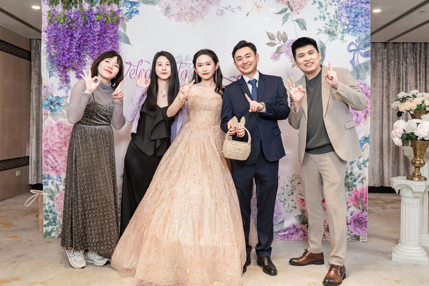 Wedding party poses in front of pink and purple floral backdrop at elegant indoor celebration venue.