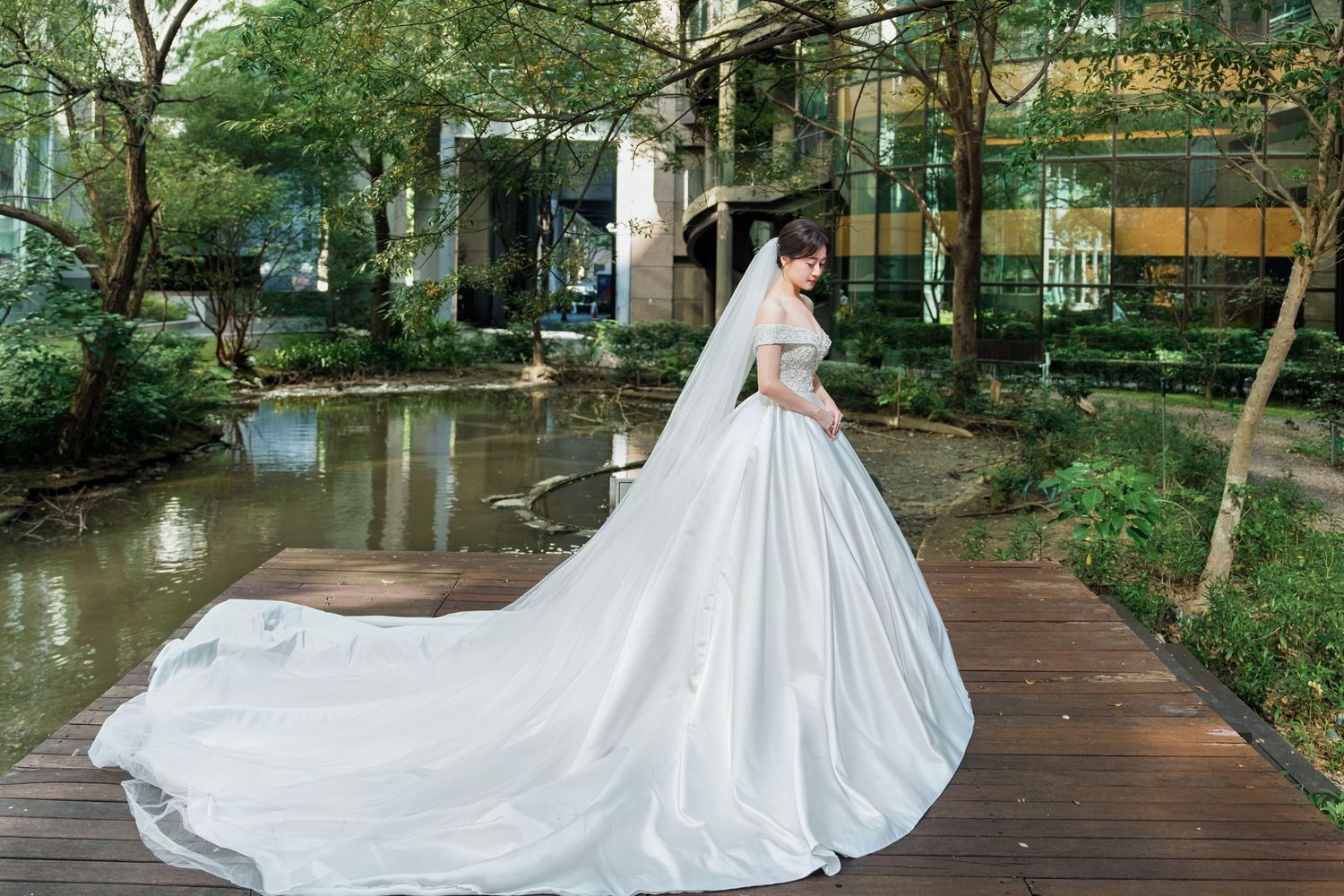 A flowing white wedding gown with long train photographed on a wooden deck near a tranquil garden pond.