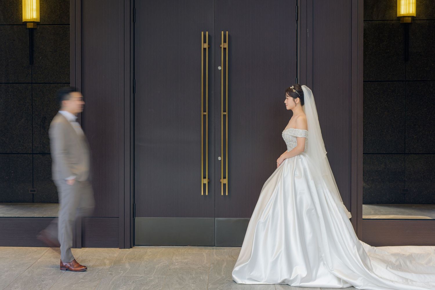 A bride in a white ball gown and veil stands by modern dark doors while a figure walks past in the background.