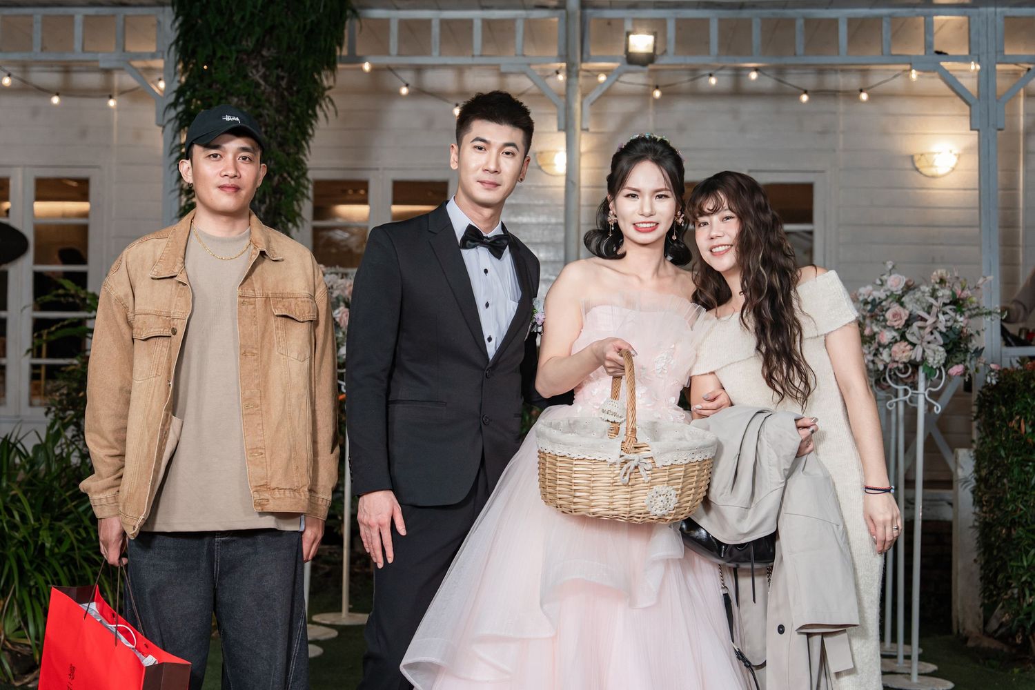 Wedding guests pose together in formal attire at an outdoor evening venue with brick walls and lighting.