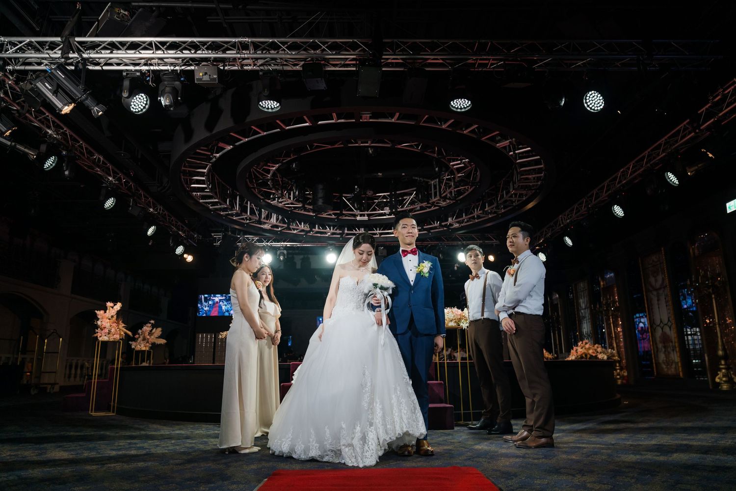 A romantic wedding ceremony takes place on a red carpet inside a rustic venue with dramatic overhead lighting and dark ambiance.