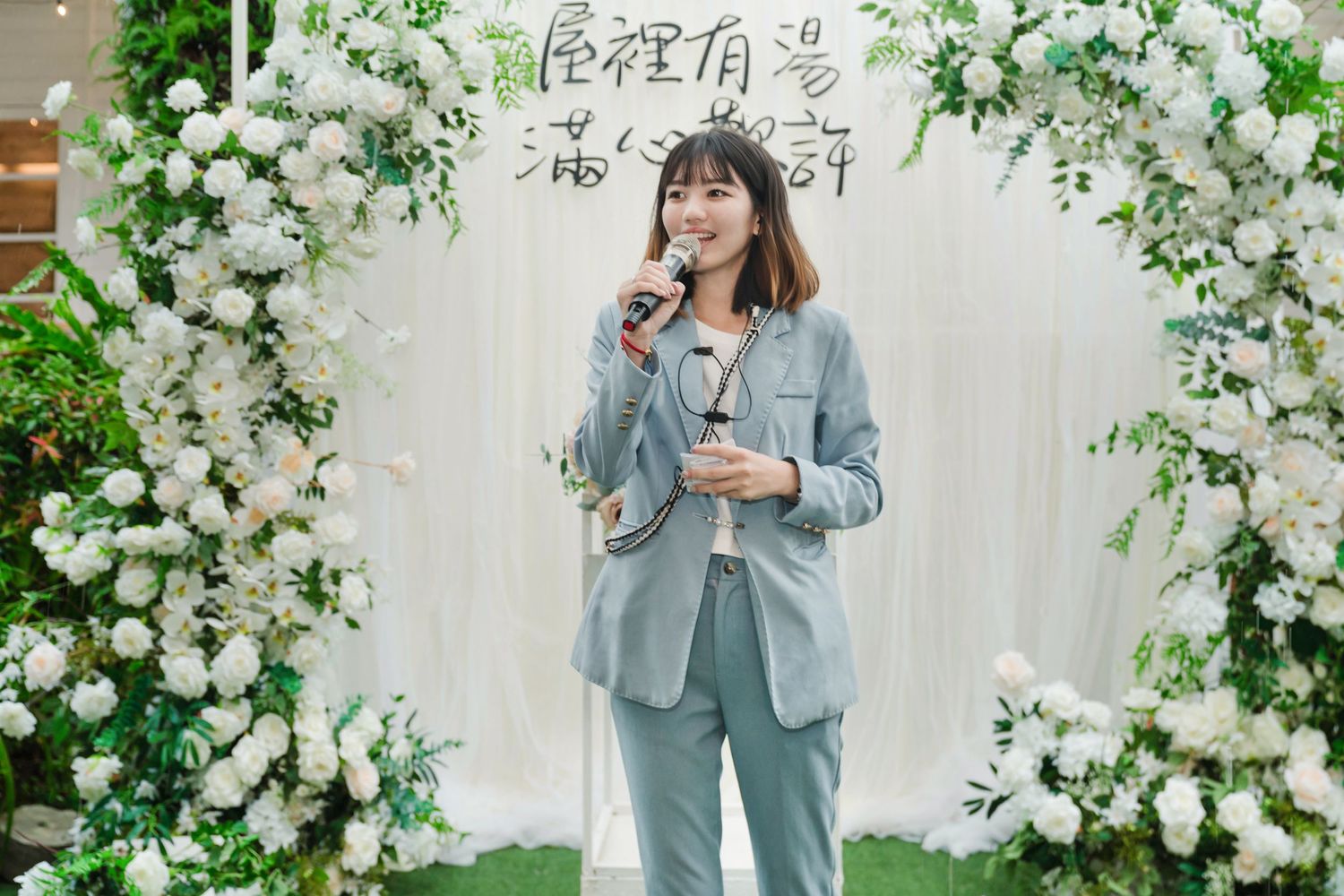 A person stands under a white floral arch wearing a light blue suit while speaking into a microphone.