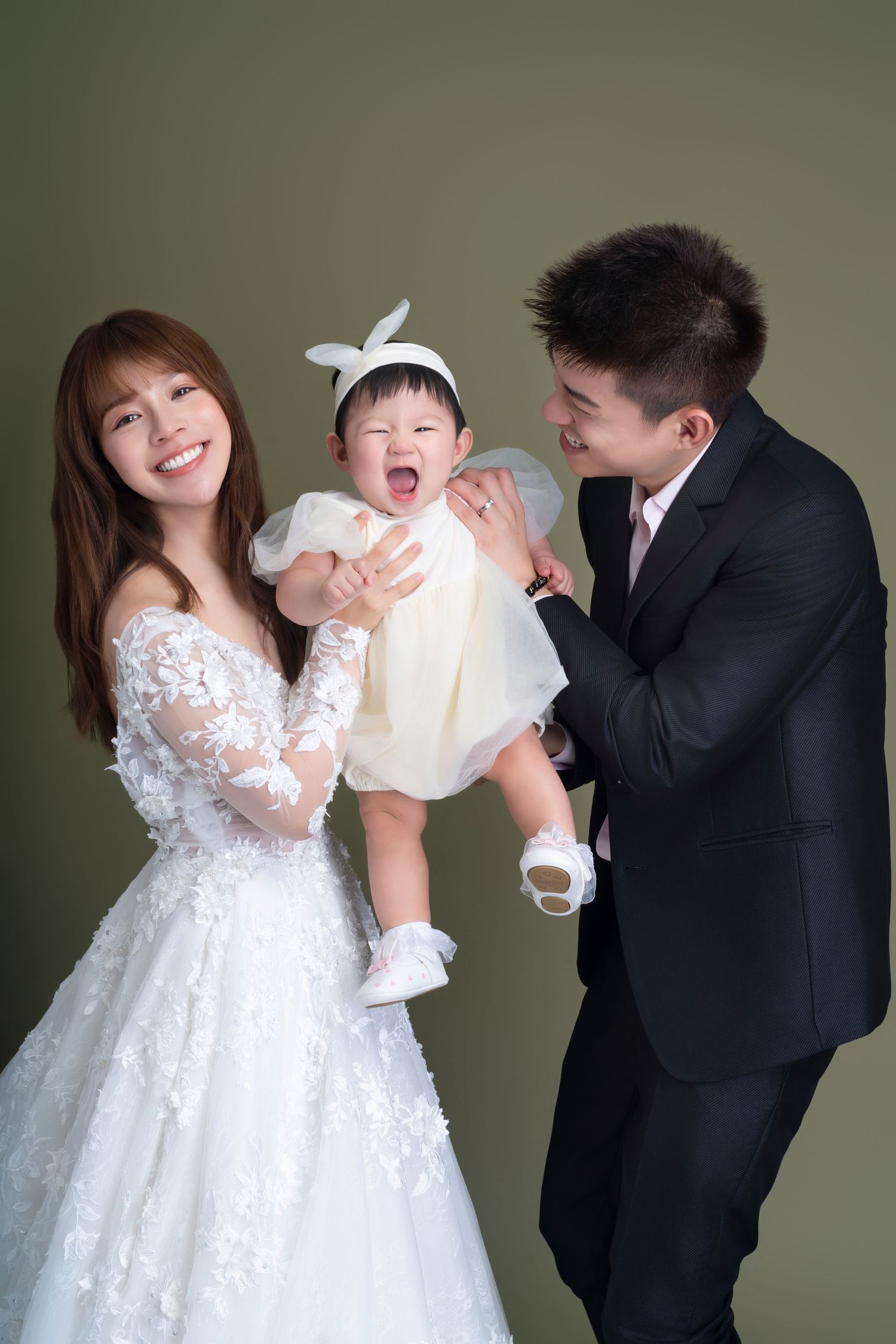Family portrait with couple in wedding attire holding a laughing baby in white dress against grey background.