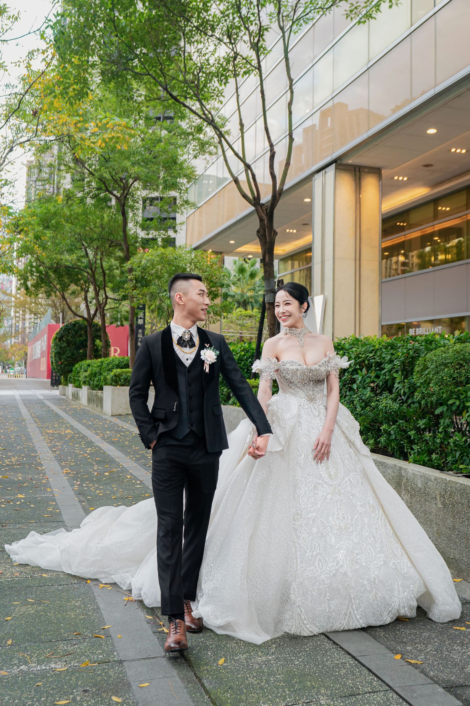 Couple in wedding attire shares playful moments on a tree-lined city sidewalk outside modern glass buildings.