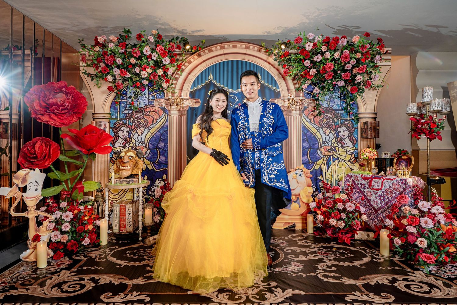 Couple in Beauty and the Beast themed wedding attire posing before floral arch backdrop with roses and decorations.
