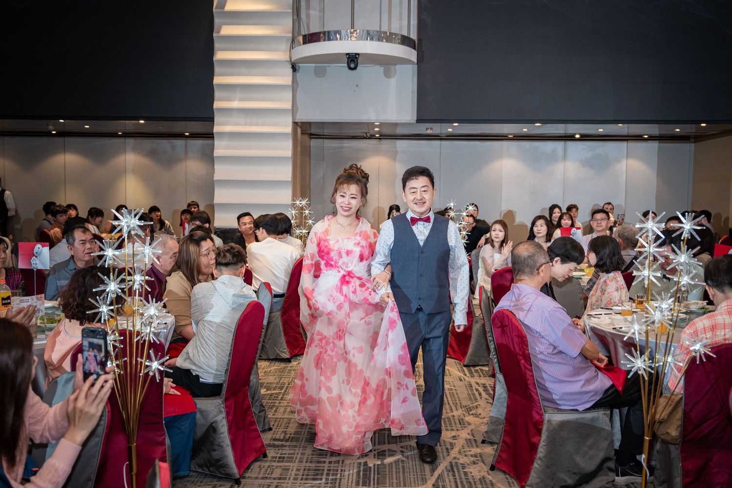 Bride in floral gown and groom in gray suit walk through decorated banquet hall during wedding reception.