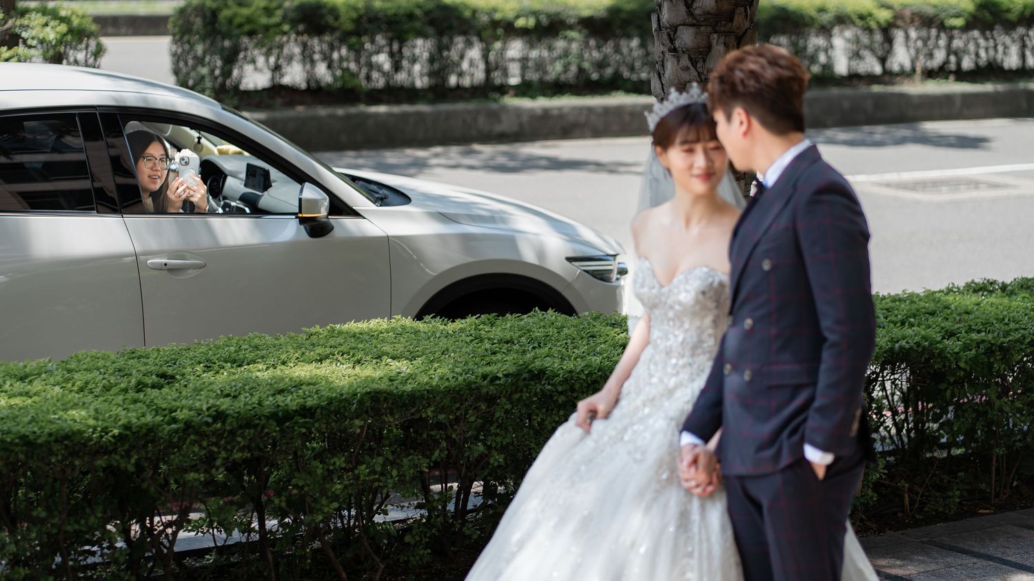 A bride in a white gown and groom in a suit walk hand in hand outdoors near a white car on a sunny day.
