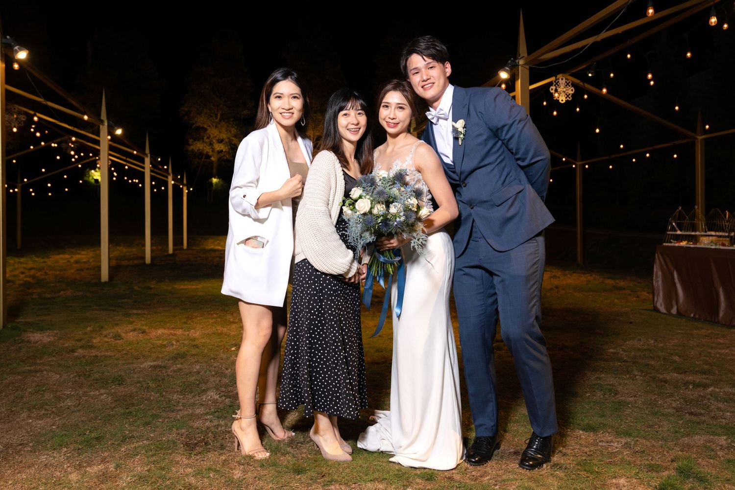 Group of wedding guests and a bride in white dress gather for an outdoor evening photo under string lights.