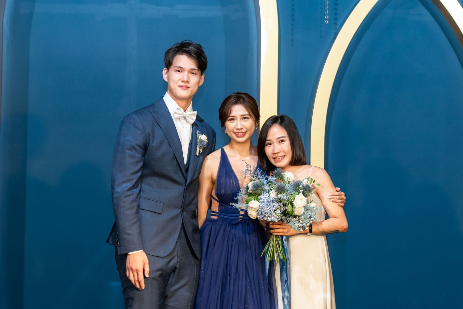 Three people in formal attire pose together against a blue wall at an elegant event, with one holding a floral bouquet.
