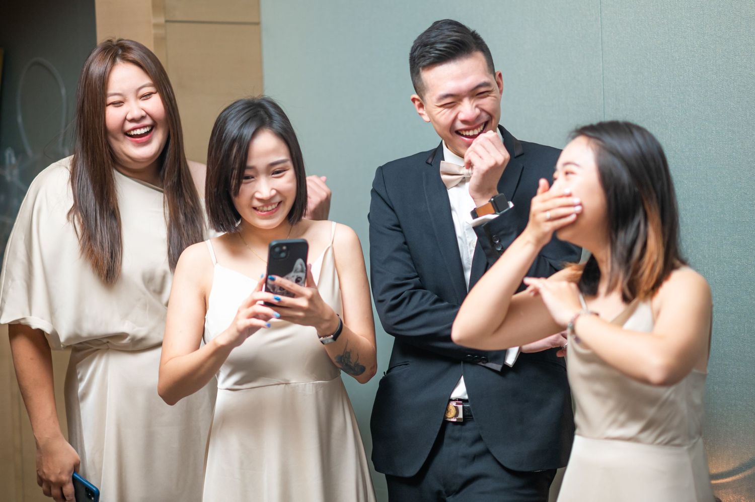 Group in formal wear laughs together while looking at a mobile phone against a gray wall.