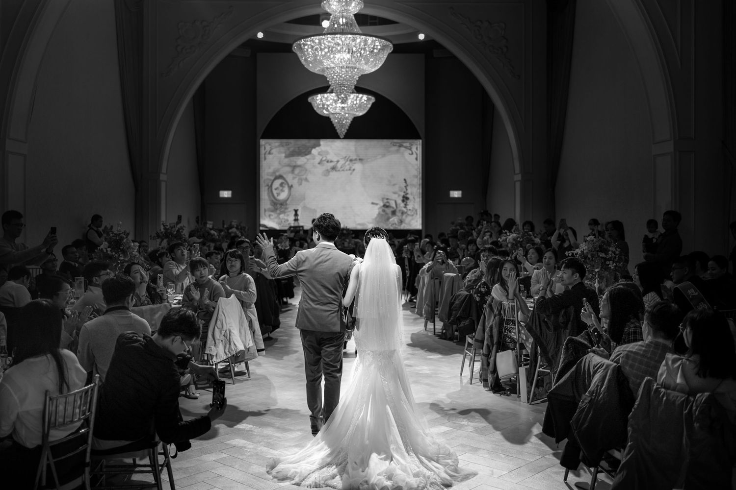 A black and white photograph of a couple walking down the aisle at their wedding ceremony with guests seated on both sides.