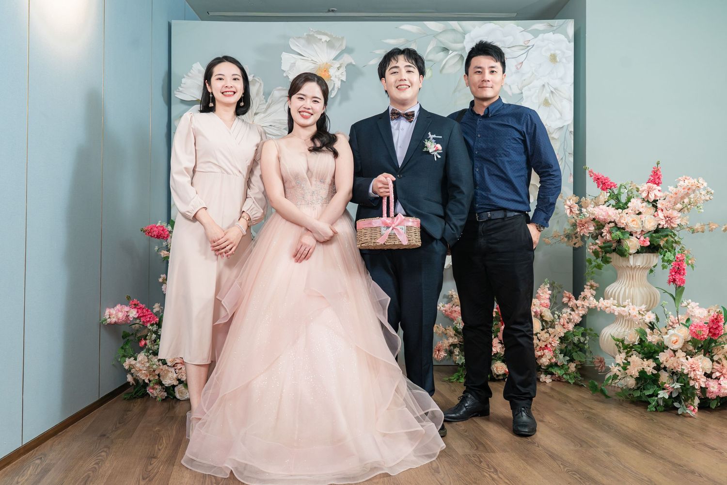 A wedding party poses in formal attire against a mint green wall with pink floral decorations.