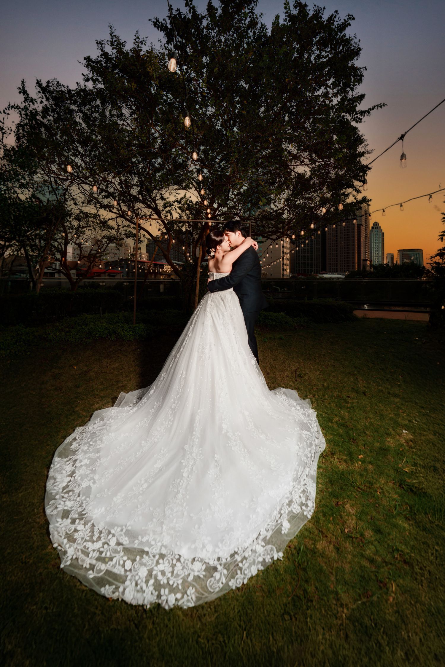 A couple in wedding attire silhouetted against a sunset sky with a dramatic lace wedding dress train on grass.