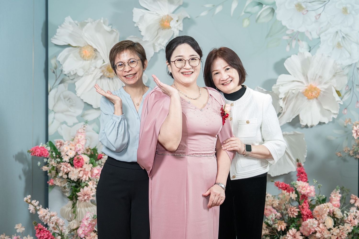 Three people pose together against a floral backdrop with pink and white flowers in a professional photo setting.