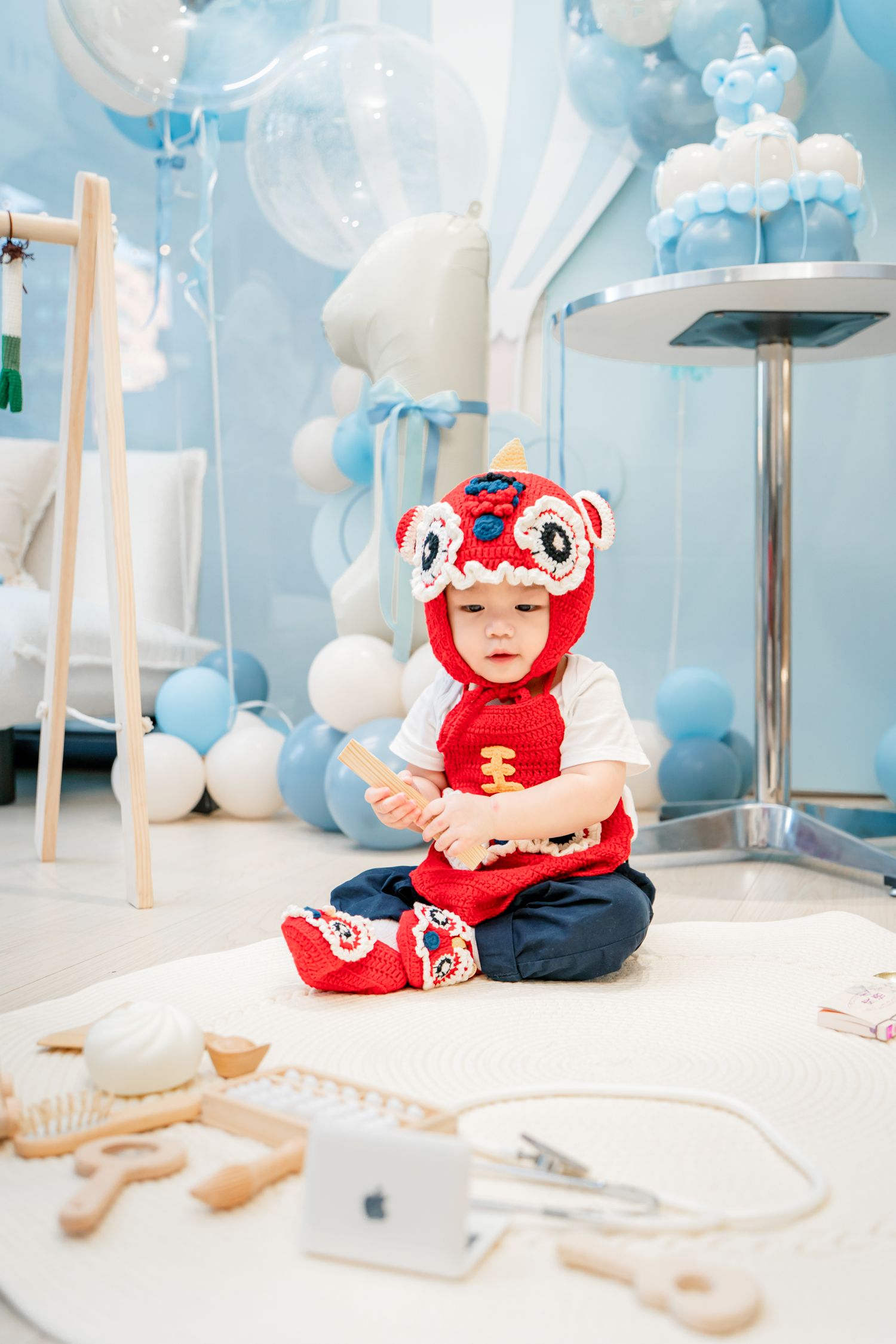 A baby in a red dragon costume sits among blue and white balloon decorations during a birthday celebration.