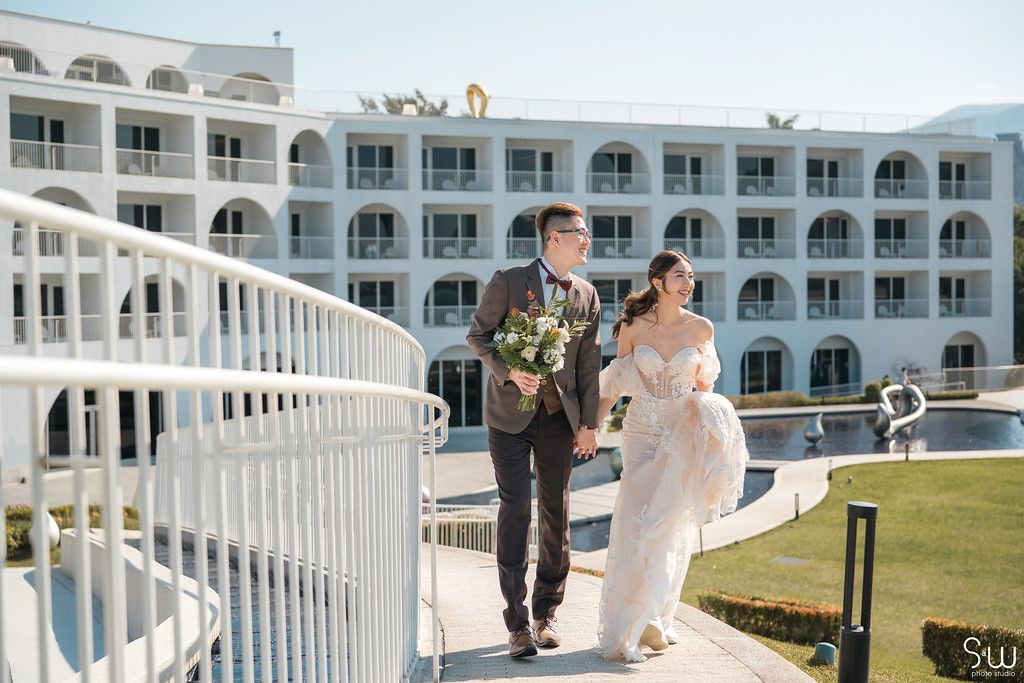 Newlyweds walk along a white balcony railing at a seaside resort hotel.