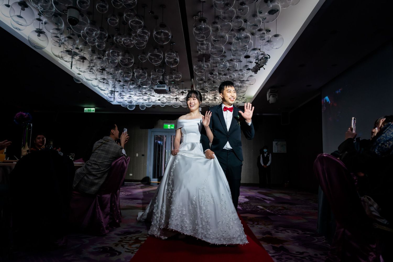 Dramatic backlit shot of newlyweds making their entrance on a red carpet under decorative lighting.