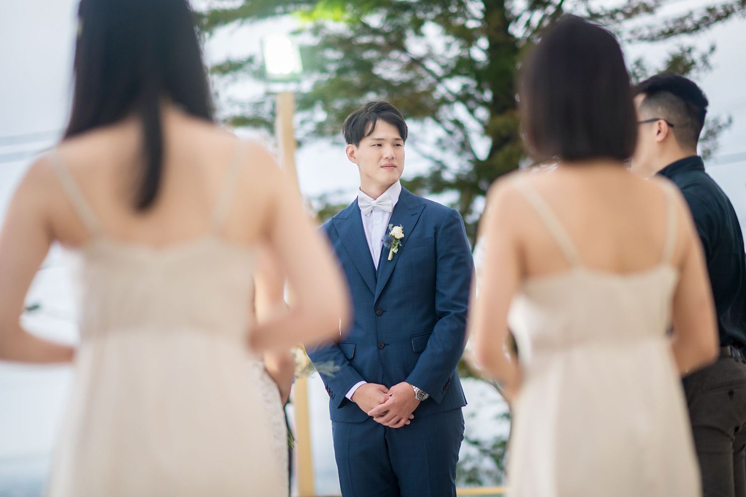 Wedding attendees in strapless blush gowns stand during an outdoor ceremony while groom in navy suit listens intently.