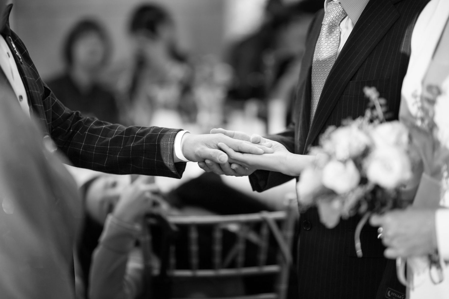 Black and white close-up of hands exchanging rings during a wedding ceremony with bouquet visible in background.