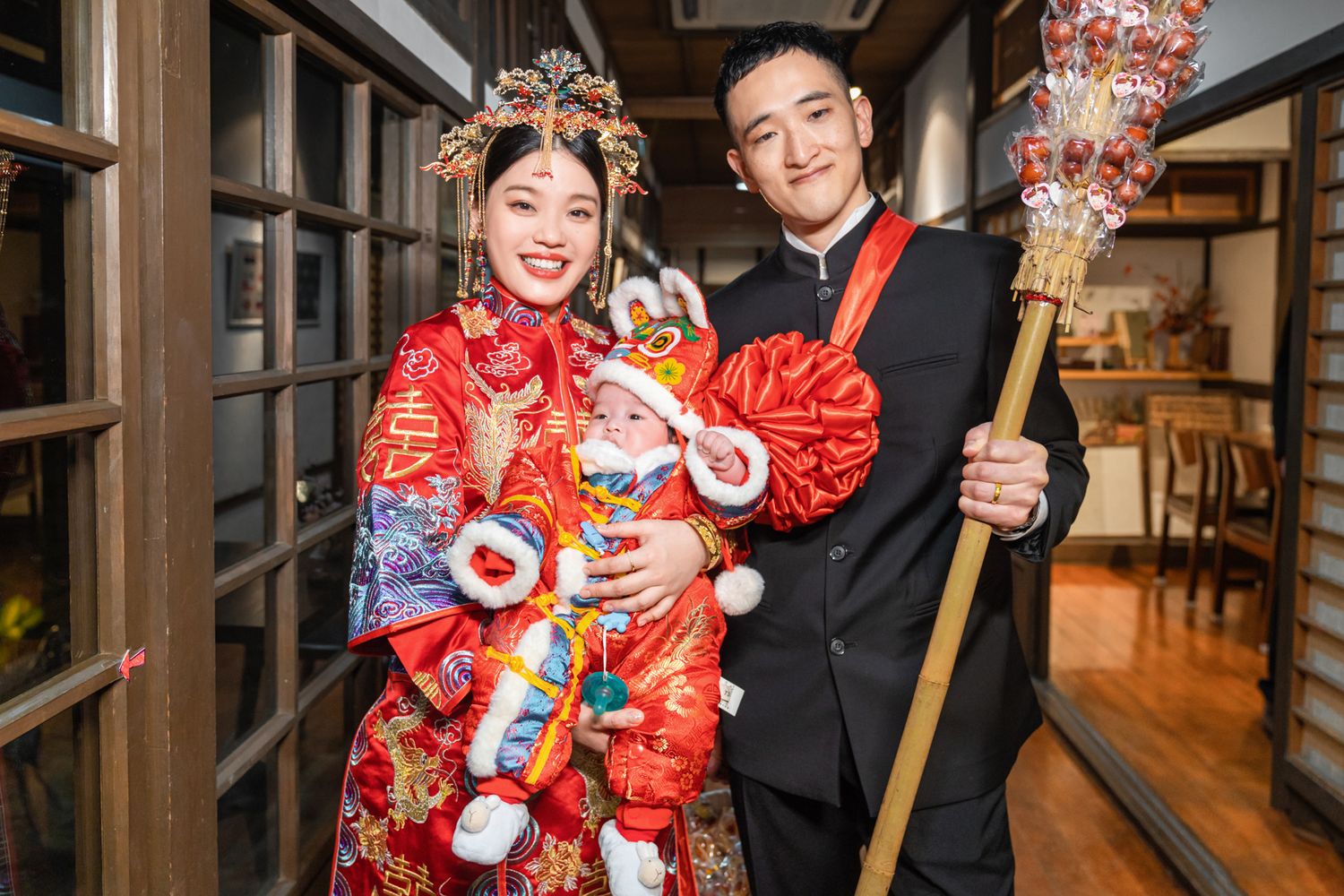 A couple in traditional Chinese wedding attire poses together in a hallway while holding decorative items and props.