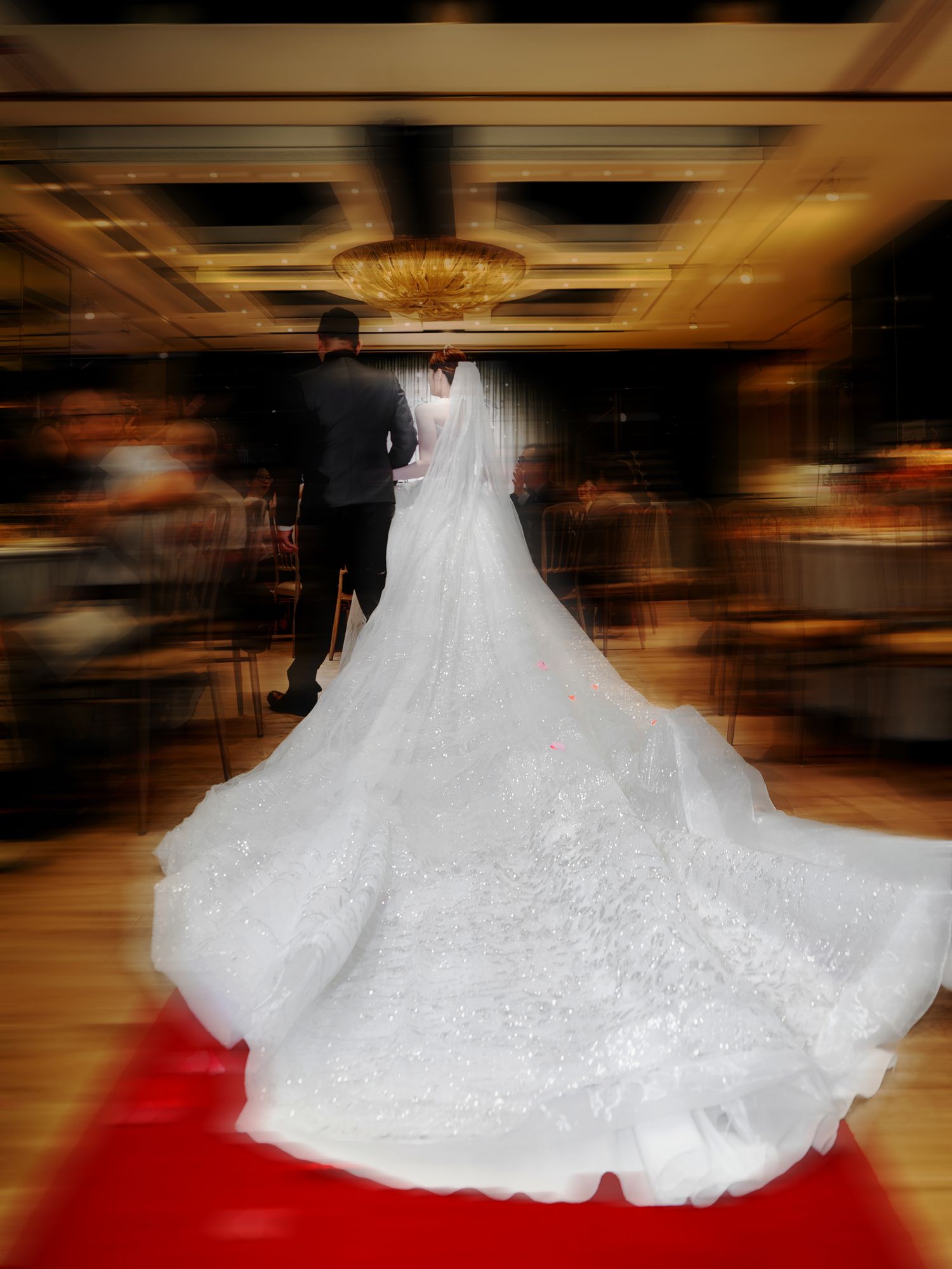 A couple walking down a red carpet in a grand ballroom with a stunning white wedding gown trailing behind.