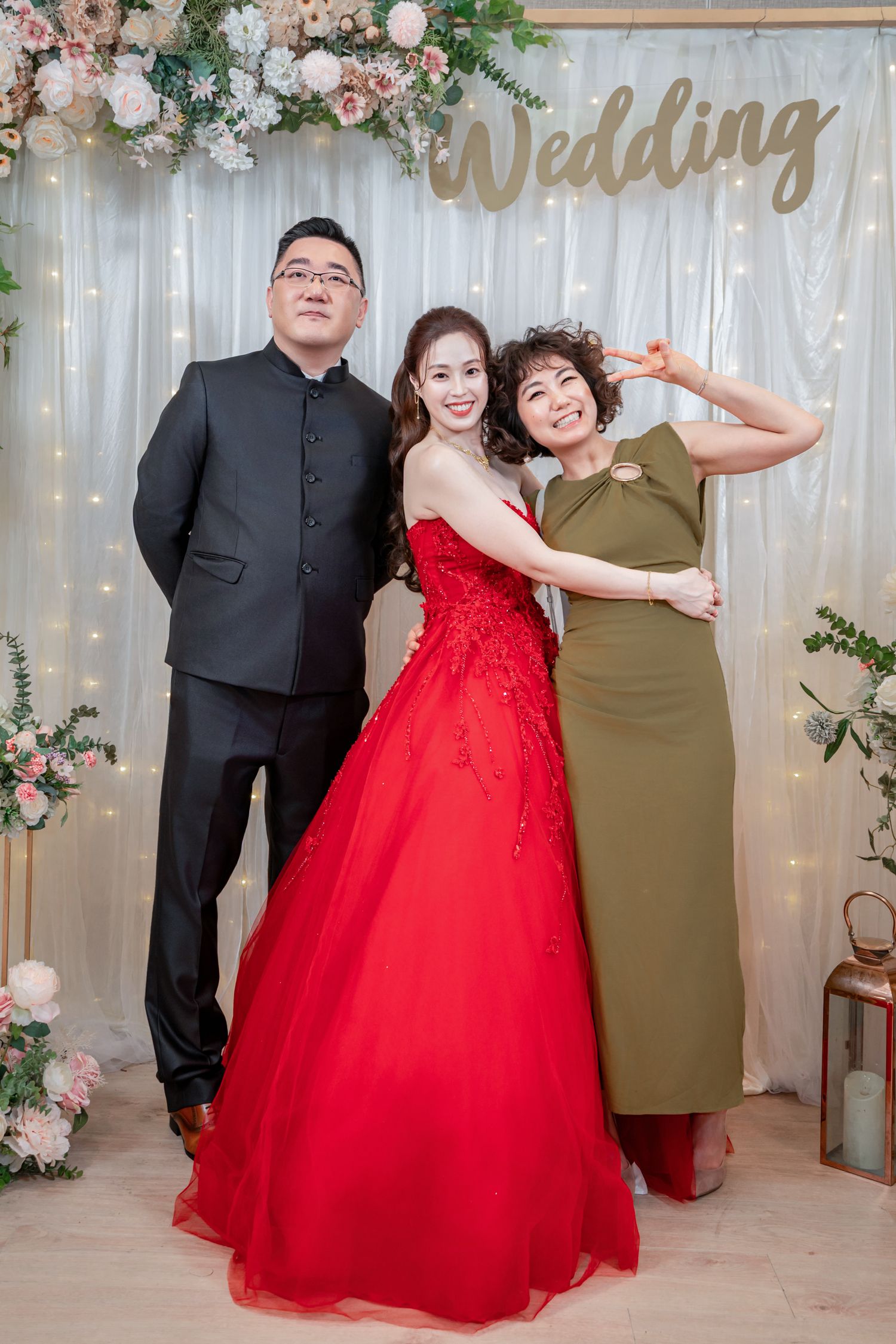 A group poses in formal attire at a wedding backdrop with floral decorations and string lights.