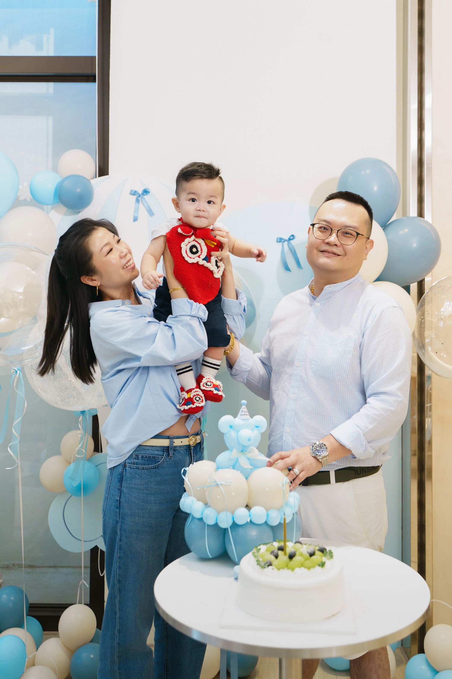 A family celebrates a baby's birthday with blue balloons and decorations in a bright modern setting.