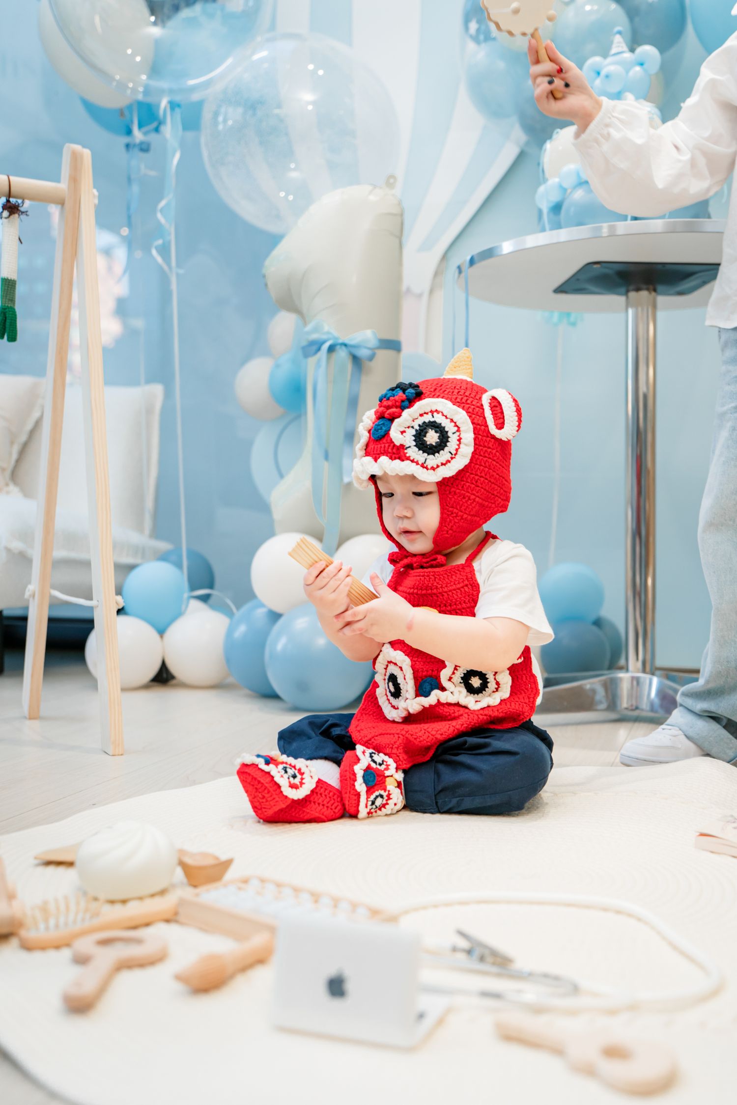 Baby in red owl costume sits on white carpet surrounded by blue and white balloon decorations at party.