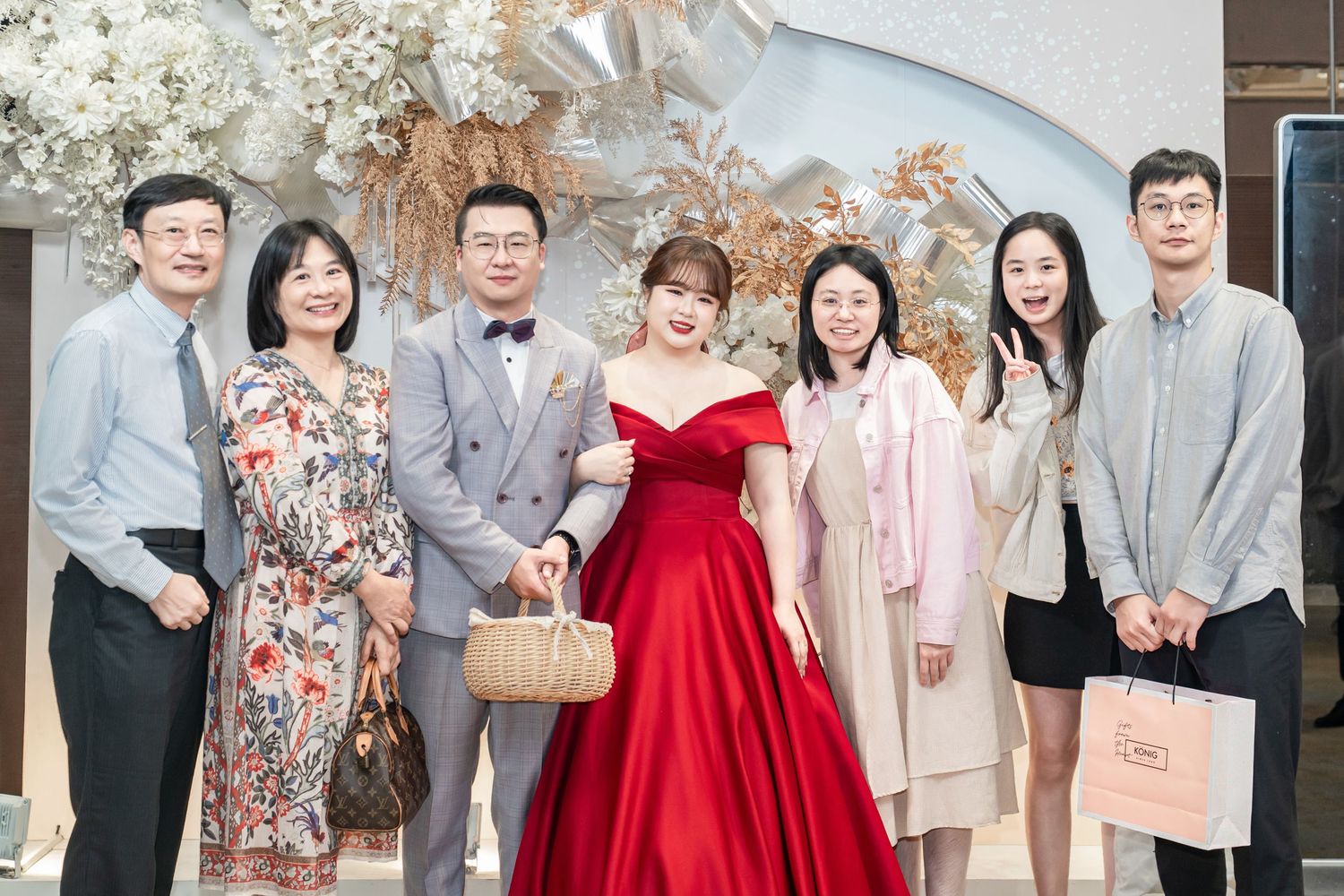A formal group photo at an event with decorative floral arrangements and a person wearing an elegant red off-shoulder gown.