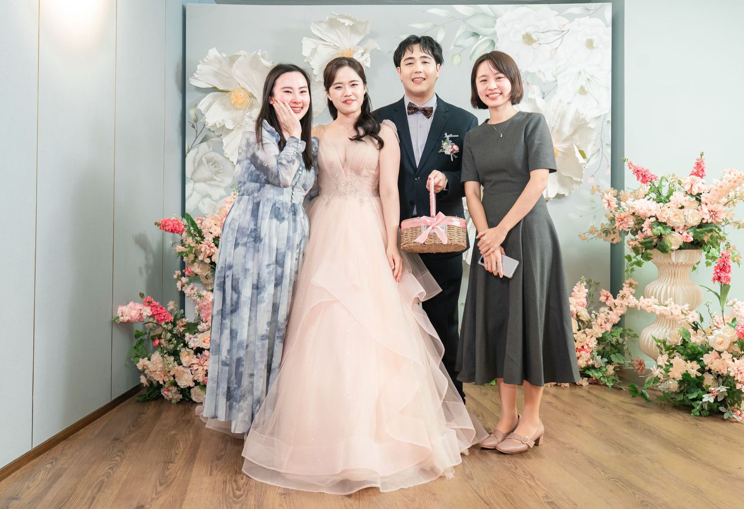 A wedding photo session with people posing in formal attire against a light backdrop with floral decorations.