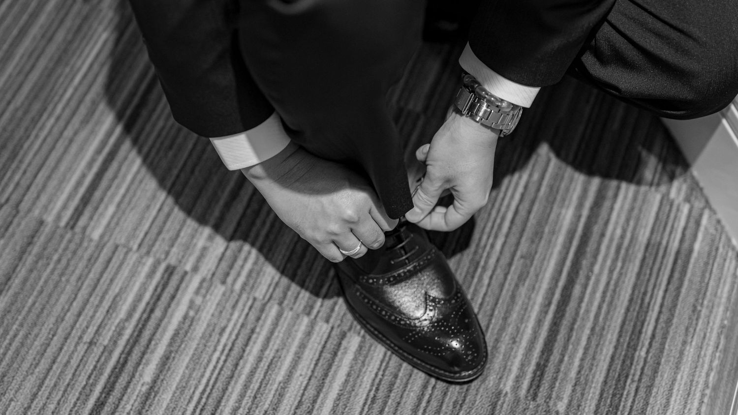 A person tying the laces of black leather dress shoes while wearing formal attire on a striped carpet.