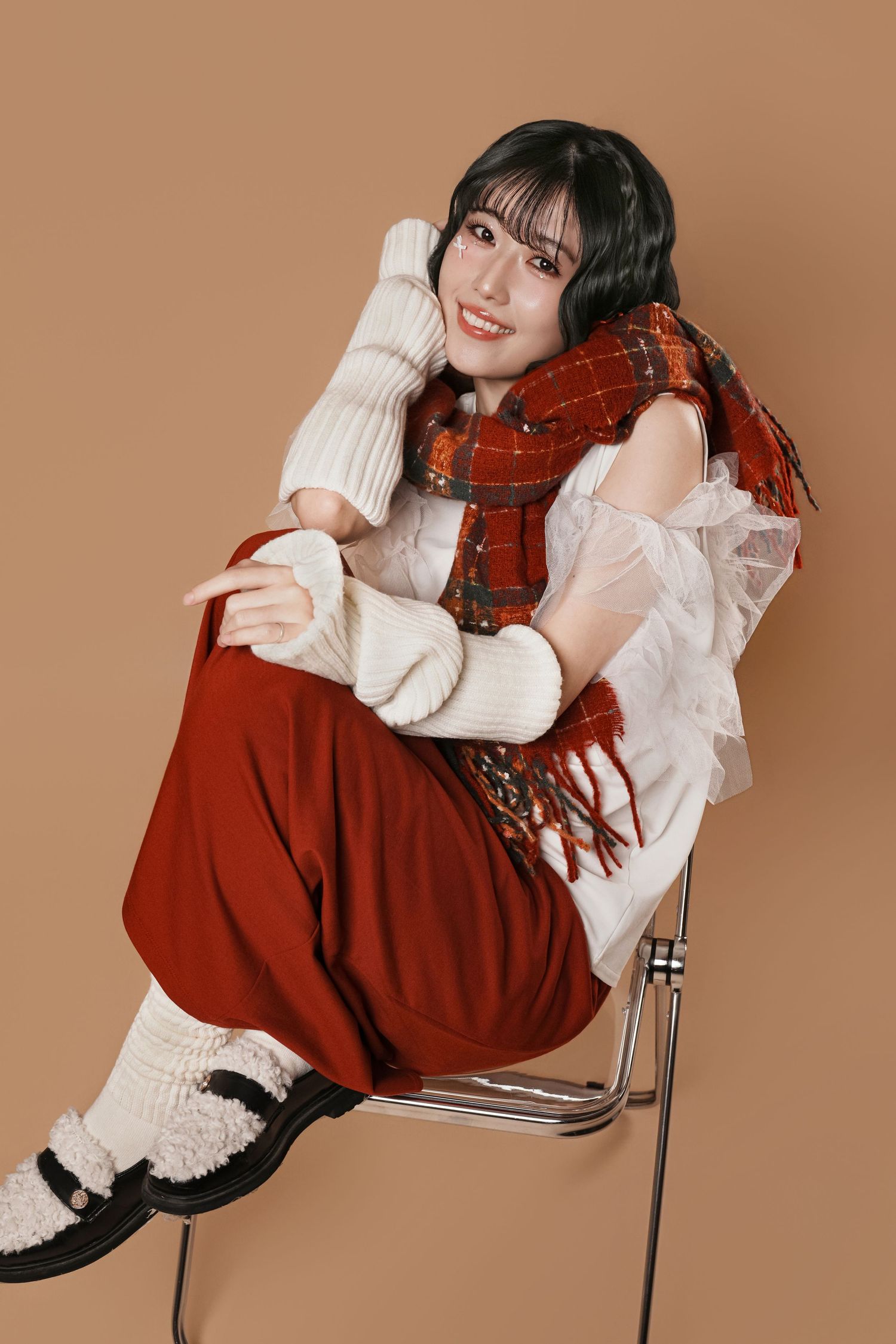Fashion model in white ruffled blouse and red scarf poses dramatically on metal chair against brown backdrop.
