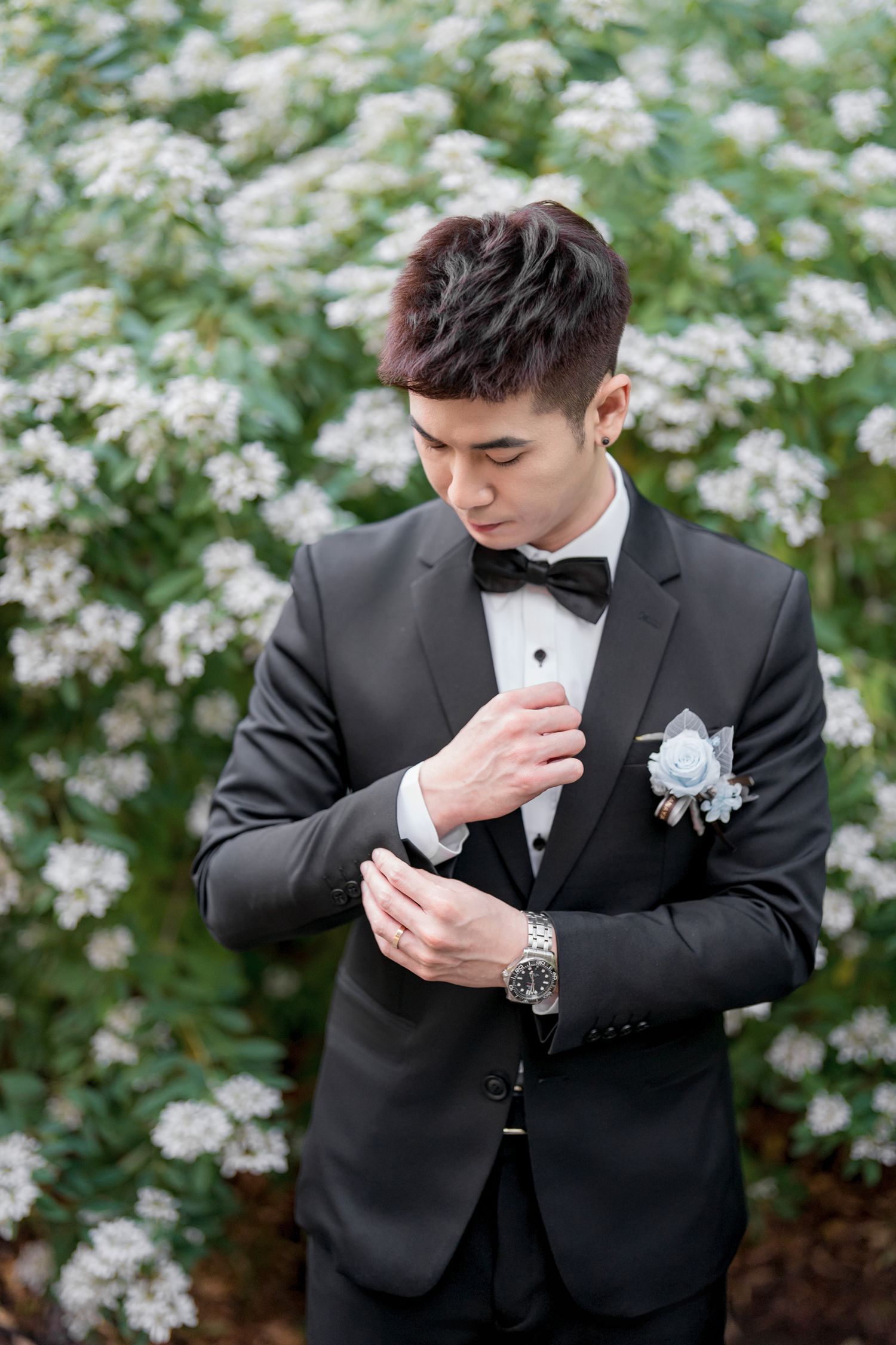 Groom in black tuxedo and bowtie adjusts his cuff against backdrop of white spring flowers.