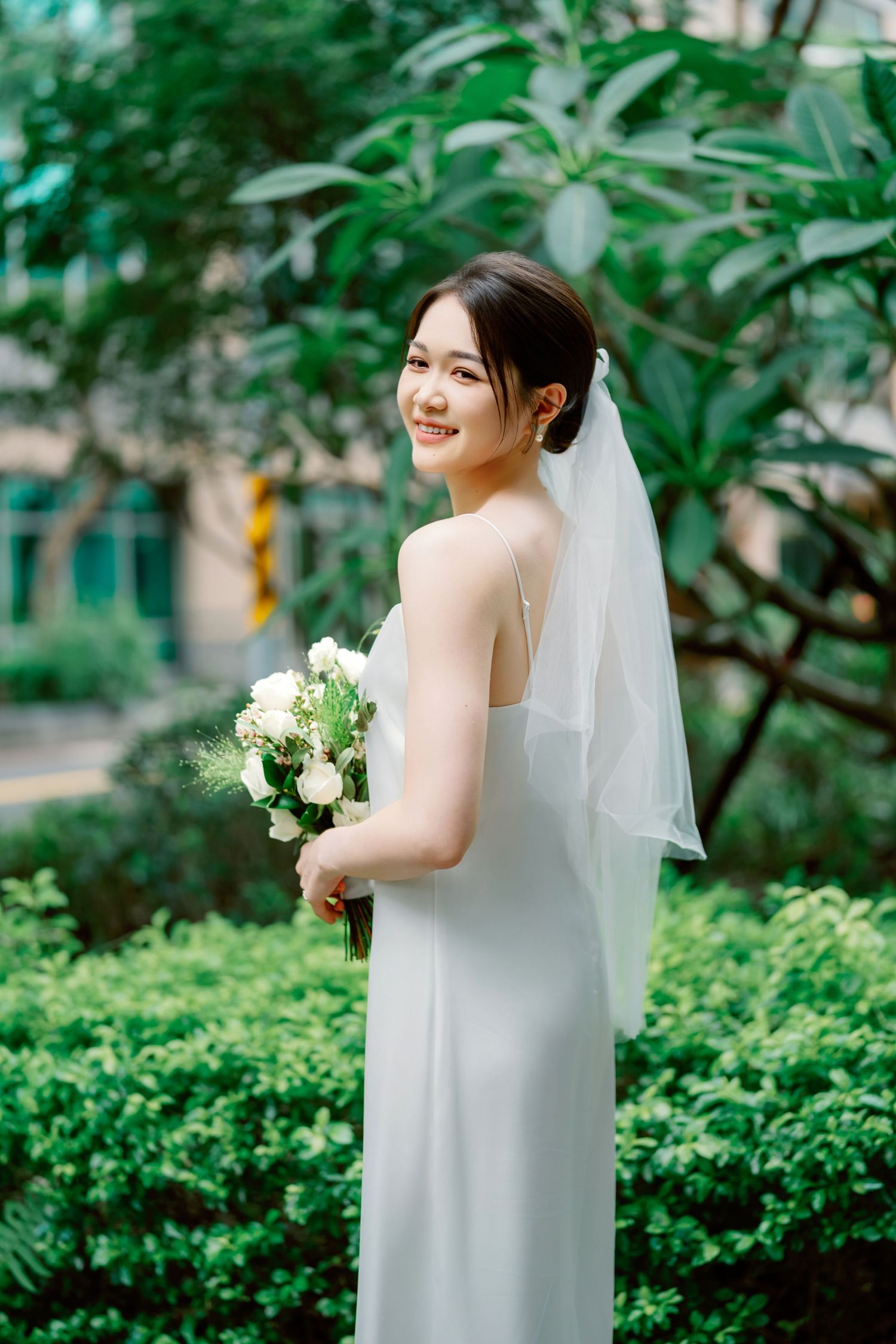 A bride in a sleek white wedding dress and veil holds a classic white bouquet while standing in a lush garden setting.