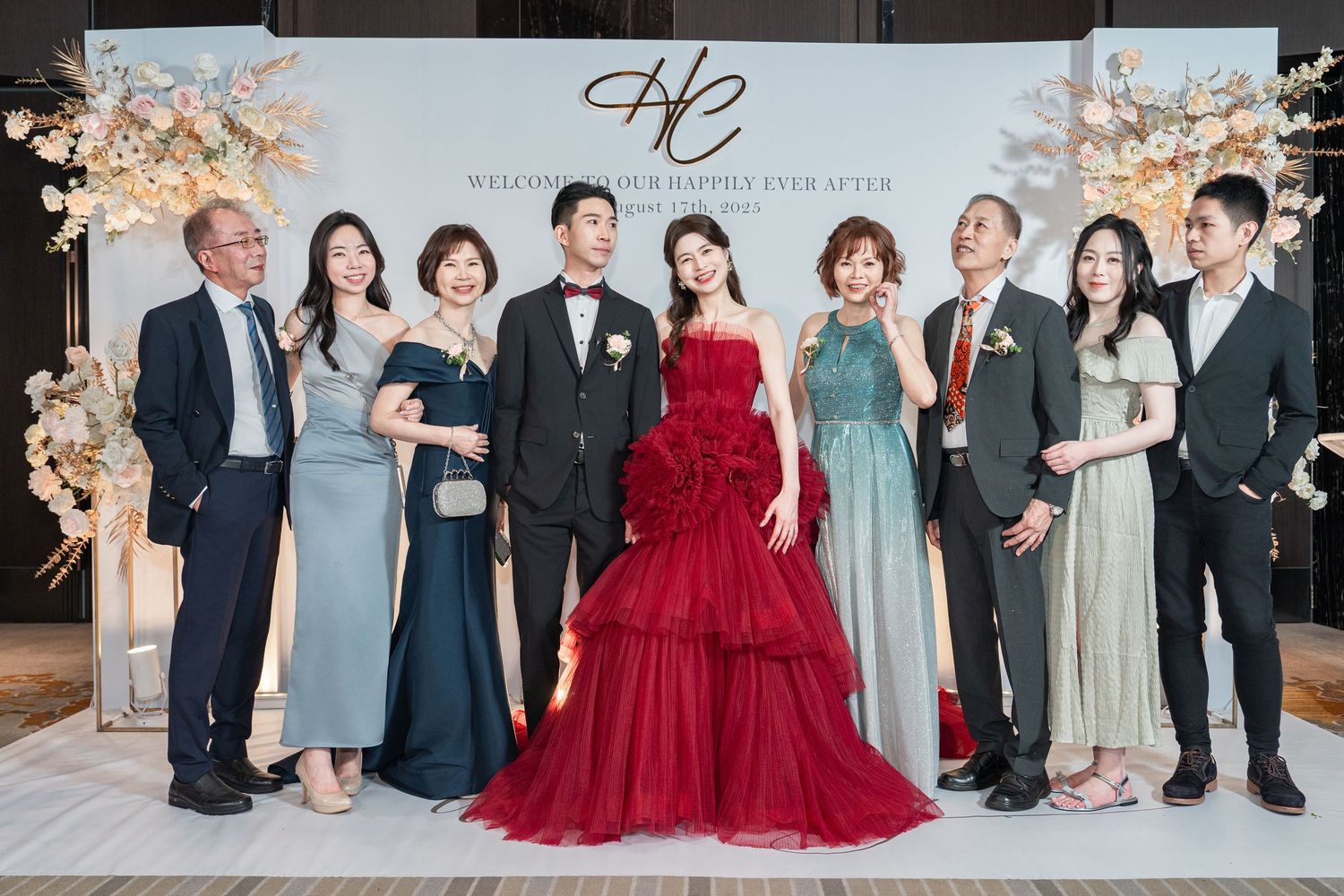 A group of wedding guests in formal attire gather around a bride in a stunning red ballgown at an elegant reception venue.