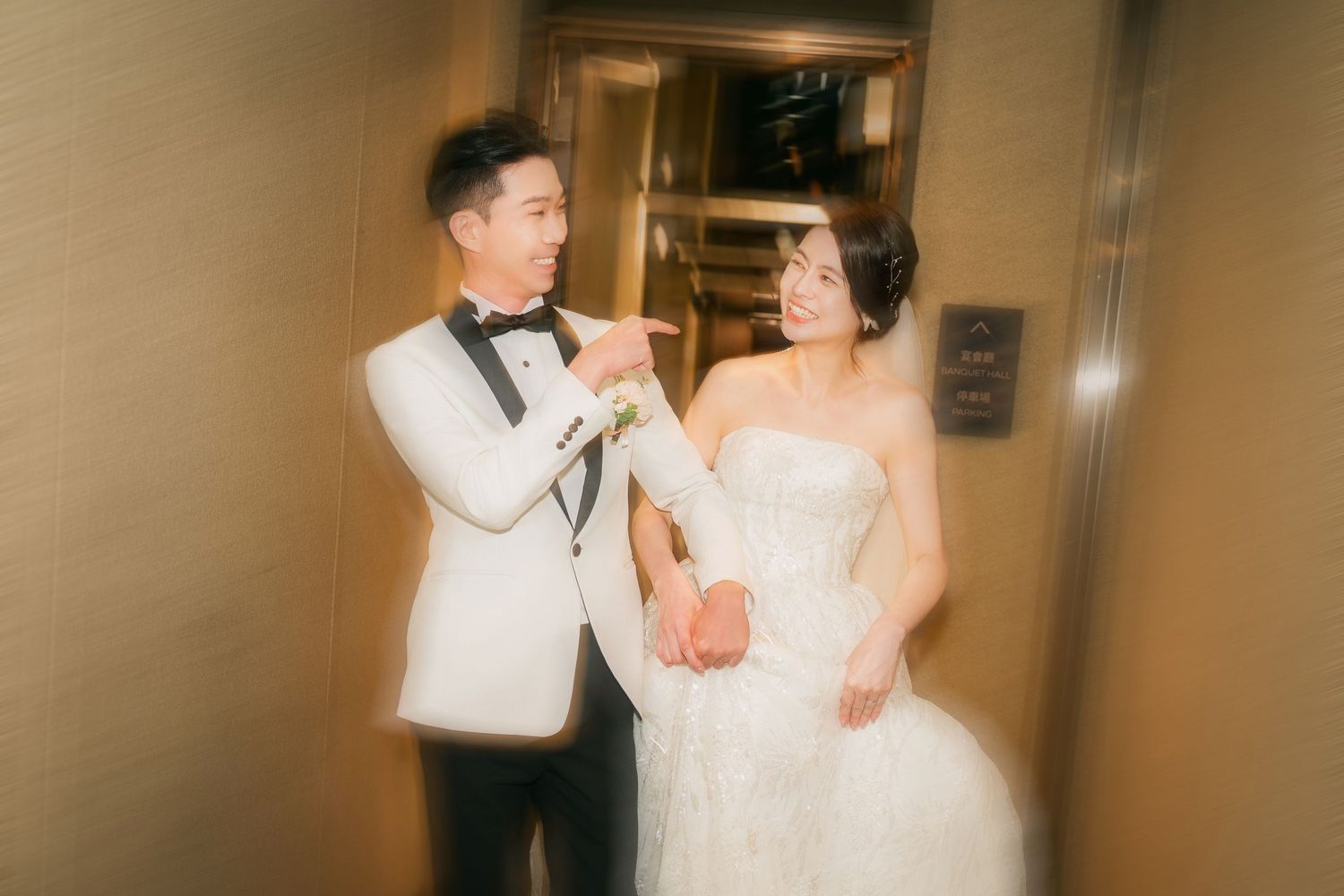 A couple in wedding attire share a joyful candid moment together in a warmly lit hallway.