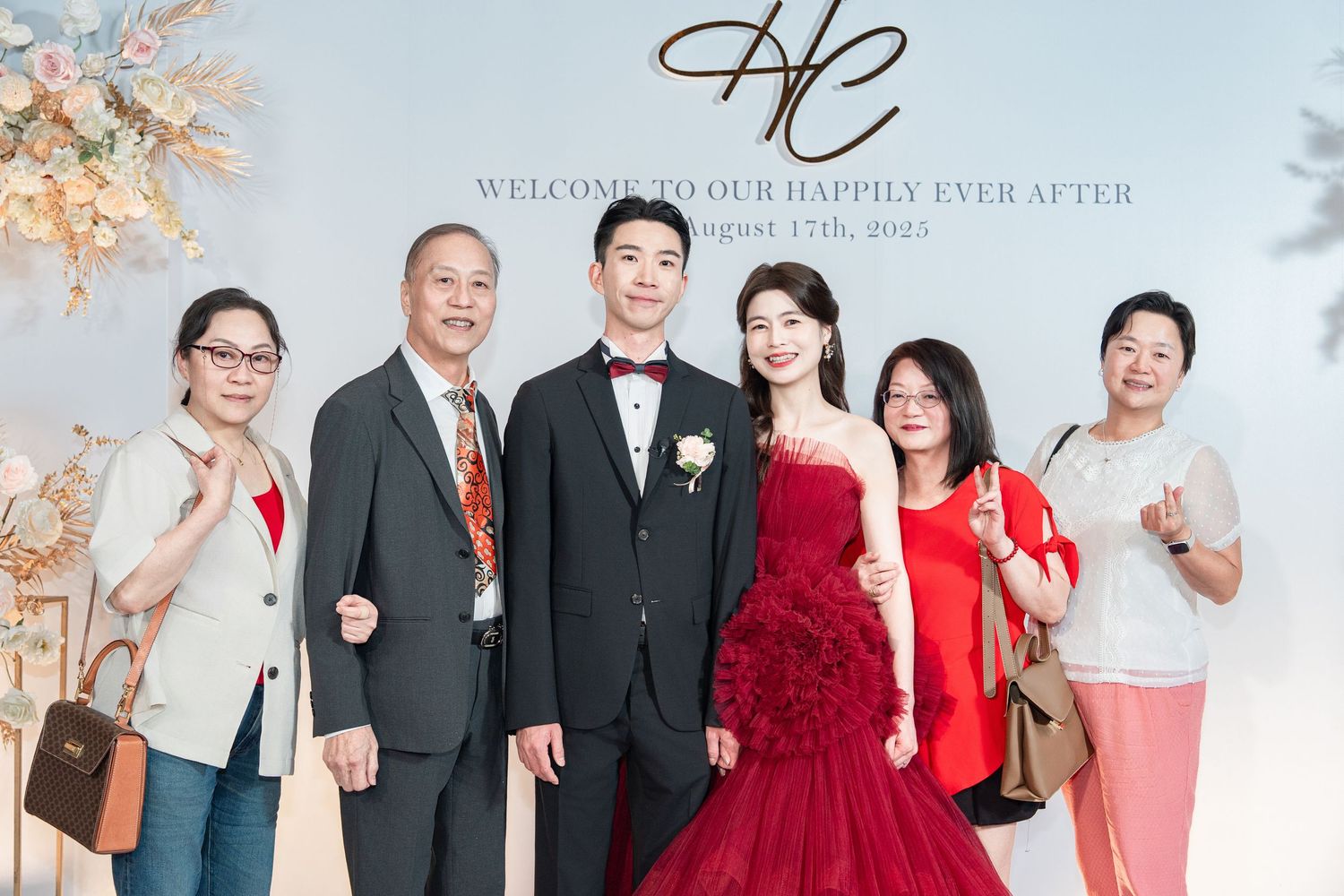Elegant wedding party in formal attire poses together with bridesmaids in vibrant red dresses against a light backdrop.