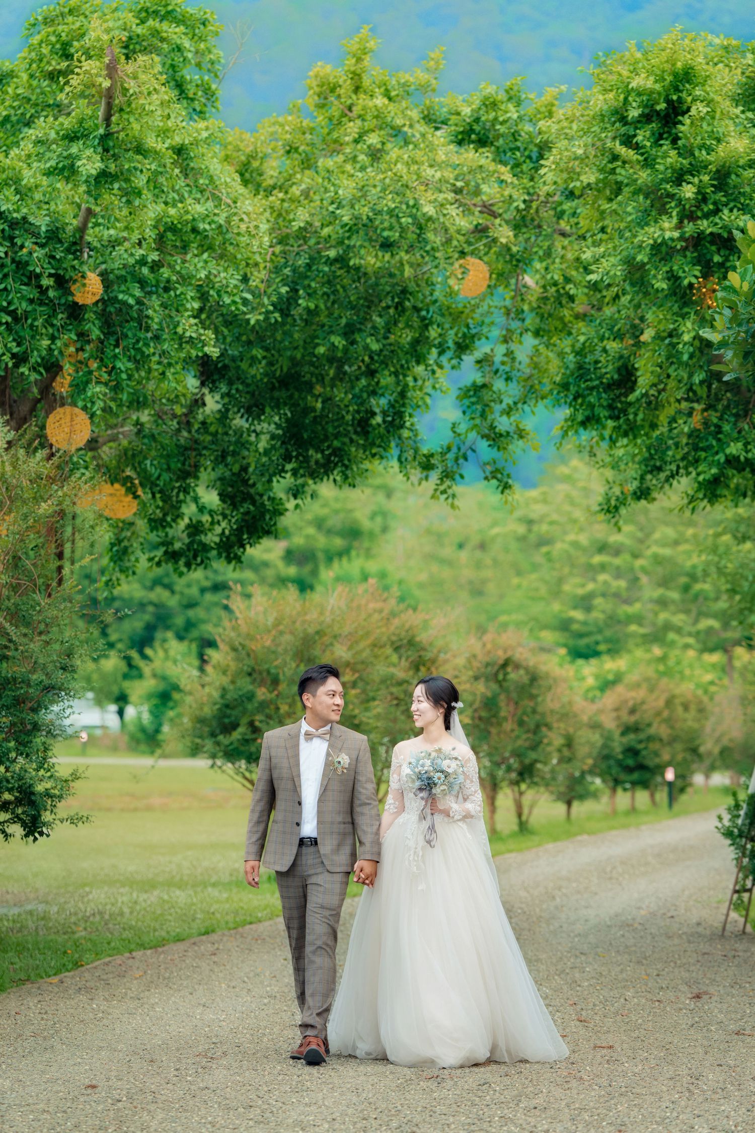 A couple in wedding attire walks along a tree-lined gravel path during a summer outdoor photo session.